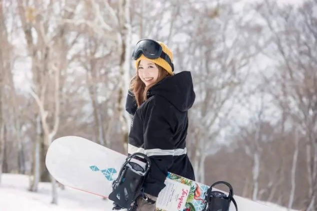 スノーボードを持って雪山で微笑む女性。冬のリゾートバイトで、仕事の合間や休日にウィンタースポーツを楽しむイメージ。