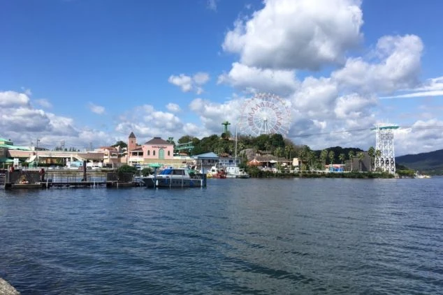 青い空に白い雲が浮かび、湖畔には大きな観覧車やロープウェイの支柱、カラフルな園内の建物が並んでいます。手前の桟橋にはボートが停泊しており、リゾート地のような景色です。