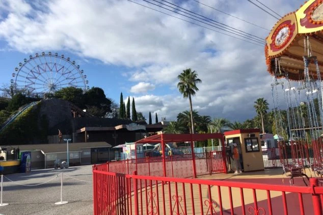 遊園地内の広場から撮影されたカット。右端には装飾の施された空中ブランコの一部が写り、遠くの丘の上には観覧車が見えます。赤い柵に囲まれた通路と、背の高いヤシの木、広々とした空が印象的な園内の様子です。