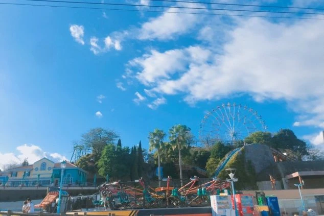 手前には回転系のアトラクションがあり、中央の丘の上には大きな観覧車とヤシの木が並んでいます。パステルカラーの建物が左側に見え、全体的に明るく開放的な雰囲気です。
