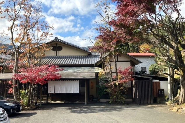 鮮やかな紅葉に囲まれた、瓦屋根と白い暖簾（のれん）が印象的な老舗旅館の外観の画像。