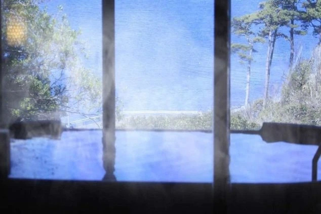 窓越しに海が見える温泉の浴槽の画像です。湯気が立ち上り、温泉の温かさが伝わります。木々も見えることから、自然に囲まれた場所でのリゾートバイトの仕事終わりに温泉で疲れを癒すイメージ