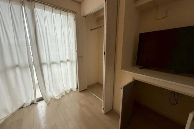 寮の居室。木目調のフローリングで、大きな窓には白いレースのカーテンがかかっている。クローゼットと、テレビを置くための収納棚が備え付けられている。