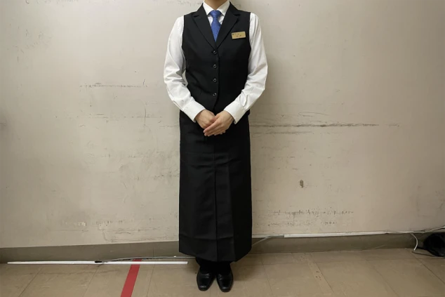 白いシャツに黒いベスト、紺色のネクタイとロングエプロンを着用。