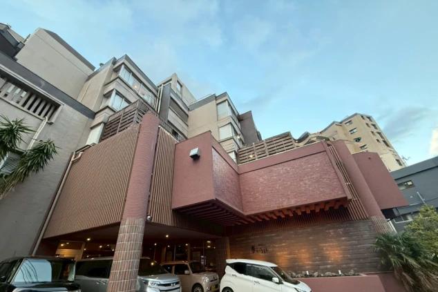 建物の外観。赤茶色の突き出したバルコニーと、背後の高層ビル。屋外の駐車場。