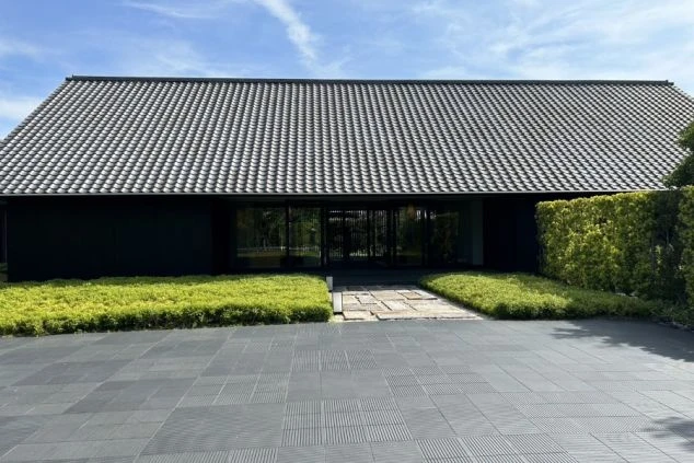 建物の正面。大きな瓦屋根と黒い壁のモダンな和風建築で、全面ガラス戸の向こうに室内が見える。手前は手入れされた芝生で、飛び石がある。