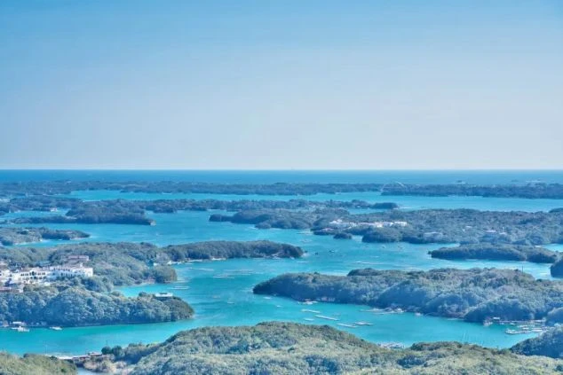 青い海に緑の島々が点在する、リアス式海岸の英虞湾（あごわん）のような多島海の景色。海面の色が鮮やかで、晴天の日の広大な風景。