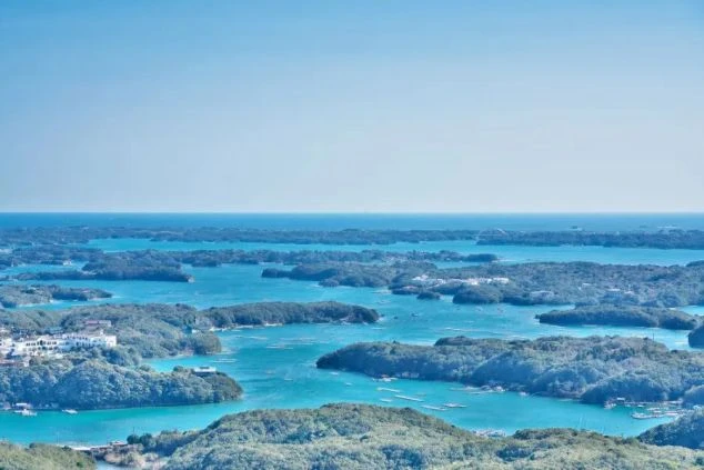 青い海に緑の島々が点在する、リアス式海岸の英虞湾（あごわん）のような多島海の景色。海面の色が鮮やかで、晴天の日の広大な風景。