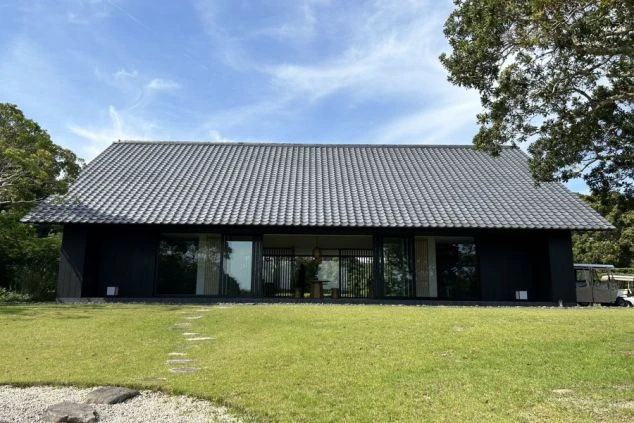 建物の正面。大きな瓦屋根と黒い壁のモダンな和風建築で、全面ガラス戸の向こうに室内が見える。手前は手入れされた芝生で、飛び石がある。