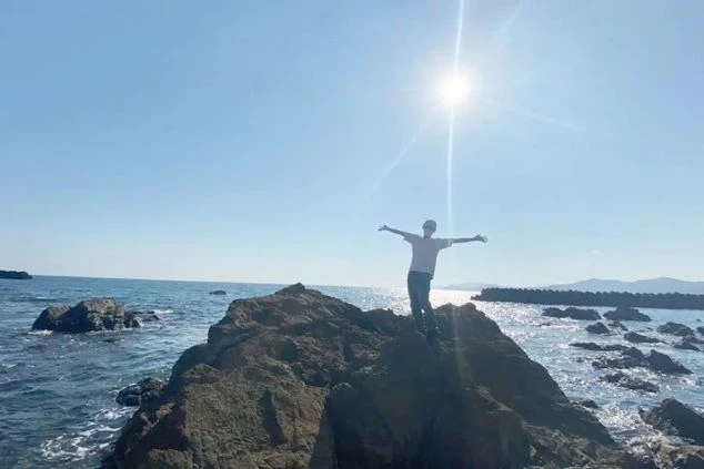 晴天の海辺で、一人の人物が大きな岩の上に立ち、両腕を広げてポーズをとっている。太陽の光が強く、海面は青く輝いている。