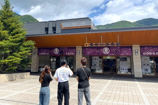 鬼怒川温泉駅のモダンな駅舎の前に立つ3人の若者。駅の看板を見上げ、これから観光を楽しむ様子。