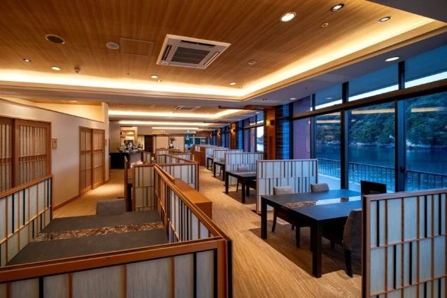 窓から海が見える、和風モダンな旅館のレストラン。段差のあるフロアにテーブル席が配置され、落ち着いた雰囲気の中でリゾートバイトのレストランサービスや配膳の仕事に興味がある方に魅力的な画像。