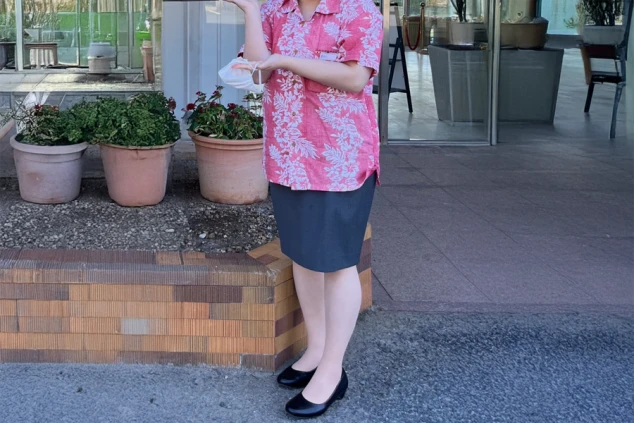 ホテルスタッフの制服を着た女性。ピンク色のアロハシャツとグレーのスカートを着用し、マスクを持っている。