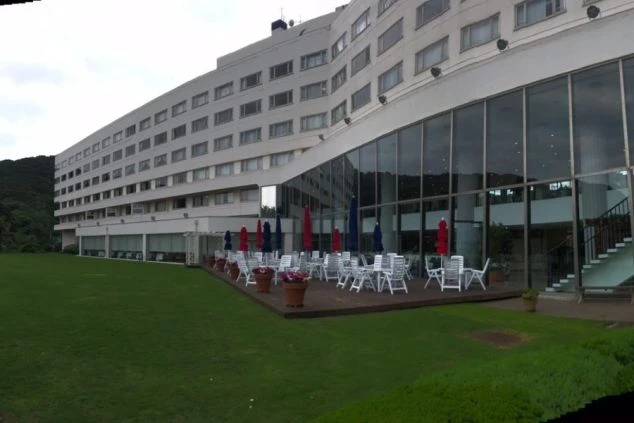 外観。白い建物と、大きなガラス窓に囲まれた低層階。手前には芝生の庭と、デッキテラスに並んだ白いテーブルと椅子、赤・青のパラソルが見える。