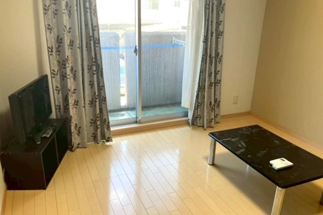 寮の居室スペース。明るいフローリングの床に、テレビ、テーブル、バルコニーに続く窓（カーテン付き）。