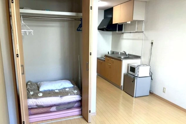 寮の室内。クローゼットに布団が収納されており、奥にはミニキッチン、電子レンジ、冷蔵庫などの設備が見える。