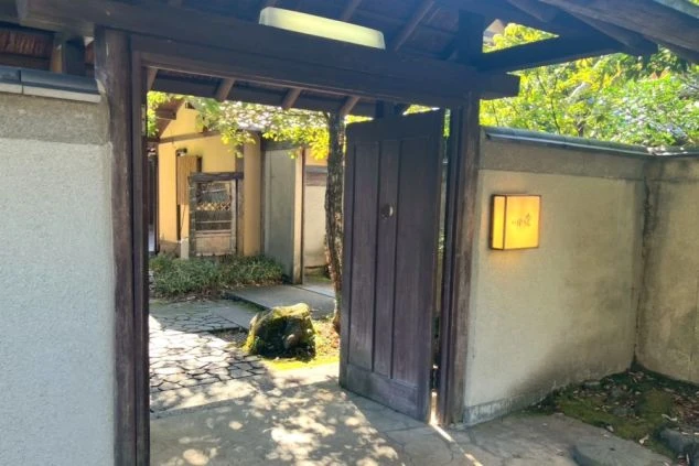木の門と瓦屋根の入り口。敷石が敷かれ、奥に和風建築の建物が見える。