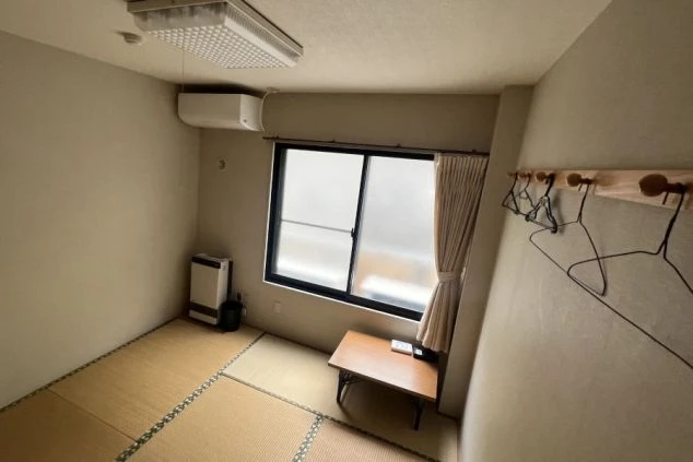 リゾートバイトの寮の部屋の中を撮影した画像。部屋には畳が敷かれており、窓際にはテーブルとヒーター、エアコンが設置されている。リゾートバイトの寮の部屋の画像。