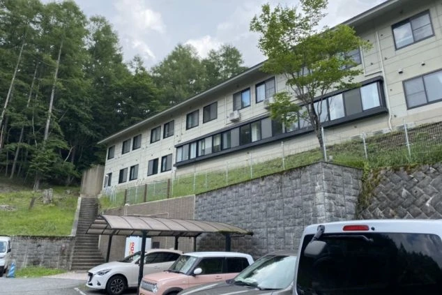 山の中に建つ、リゾートバイトの寮の外観の画像。白い壁の建物は、駐車場と階段を挟んで木々に囲まれている。リゾートバイトの寮の外観の画像。