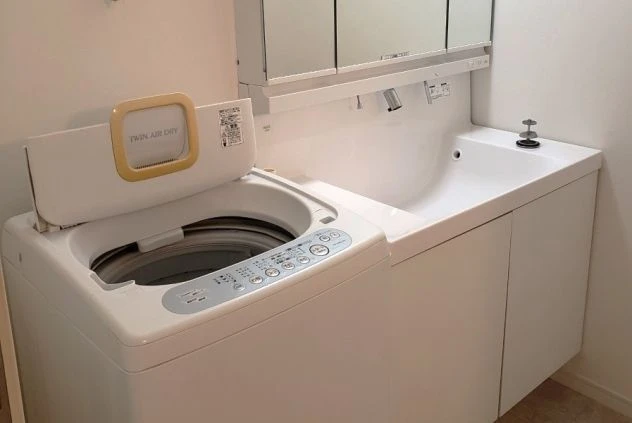 白い洗面台の横に、蓋が開いた状態の白い縦型洗濯機が設置されている、清潔感のあるサニタリースペース。