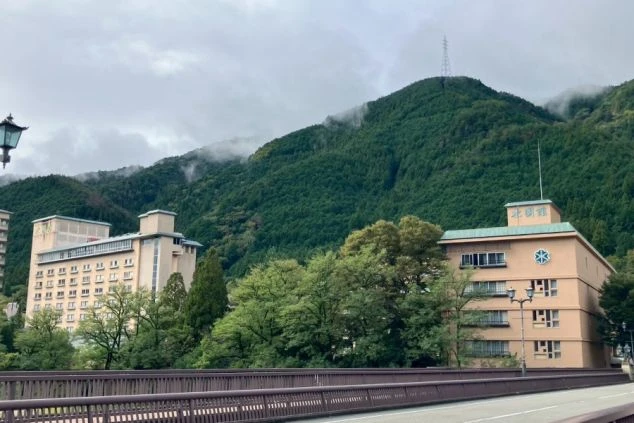 山と森に囲まれた、川向こうに建つベージュ色の水明館の建物群の外観。手前の建物には「北館」の文字が見える。