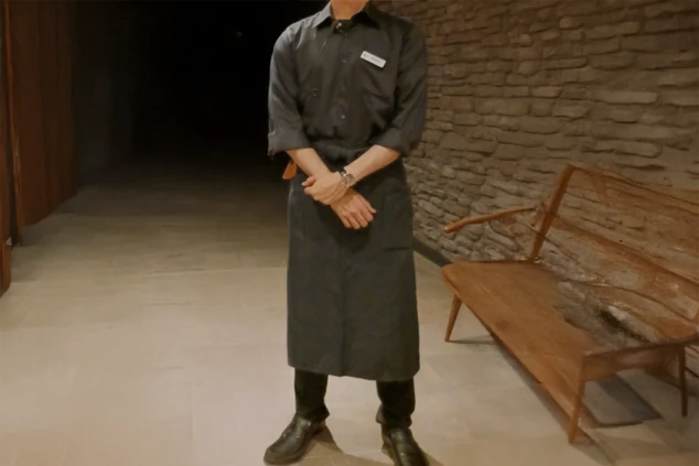 リゾートホテルのレストランで働く男性スタッフ。濃いグレーのシャツとエプロンを着用。落ち着いた雰囲気の制服で、レストランサービスやホール業務の仕事をイメージさせる。