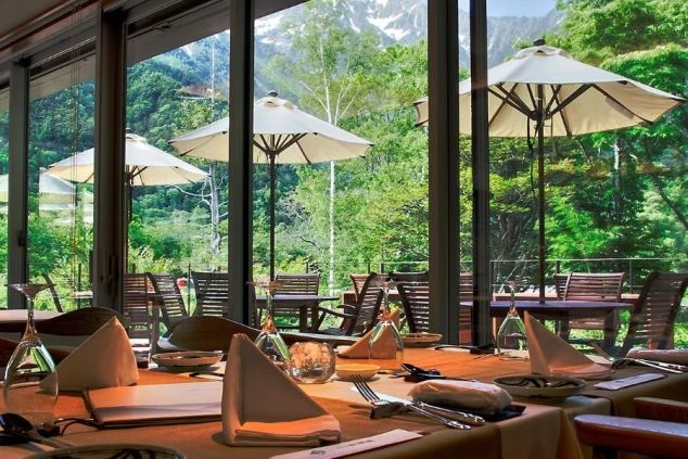 大きな窓から残雪の北アルプスと瑞々しい新緑を一望できるレストラン。テーブルにはカトラリーが整然と並び、屋外テラスにはパラソル付きの席が設置された、開放感あふれる上質な食事空間。