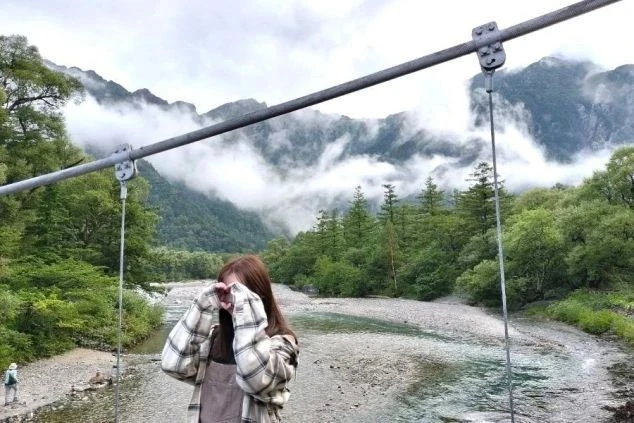 霧が立ち込める幻想的な山々を背に、吊り橋の上で手でハートマークを作る女性。雄大な自然の中で旅を楽しむ、リラックスしたひとときを切り取ったスナップ写真。