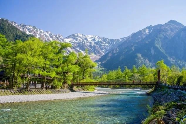 抜けるような青空と残雪の北アルプスを背景に、梓川に架かる河童橋を望む絶景。澄んだエメラルドグリーンの清流と、周囲を彩る瑞々しい新緑が調和した上高地を象徴する風景。