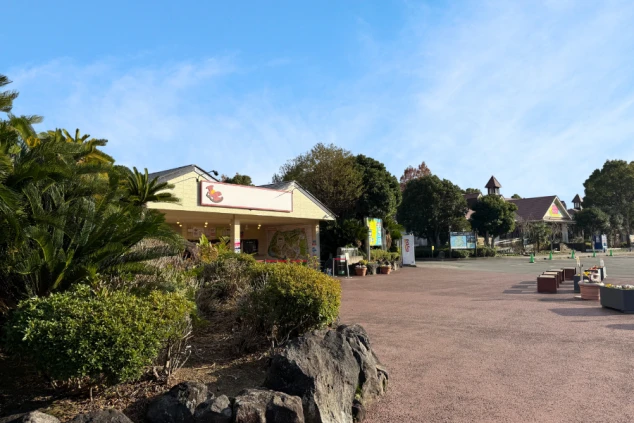 青空の下に建つ伊豆ぐらんぱる公園のチケット売り場。黄色の平屋建ての建物で、周囲にはヤシの木などの豊かな植栽があります。