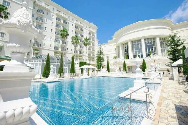 ヨーロッパの宮殿のような白い建物に囲まれた豪華な屋外プール。プールの底は青いモザイクタイルで装飾され、周囲には白い植木鉢やコニファー、ヤシの木が並んでいる。