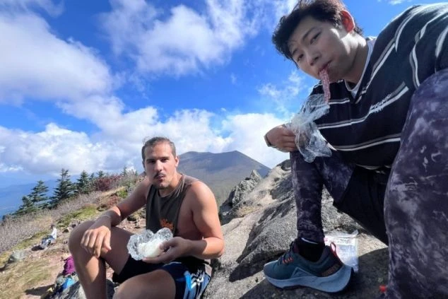 2人の男性ワクトリスタッフが山の上で座りながら、食べ物を食べている画像。リゾートバイトを通じてできた友人たちと、休日に登山を楽しんでいる様子が伝わるワクトリスタッフが撮影したオリジナル画像。