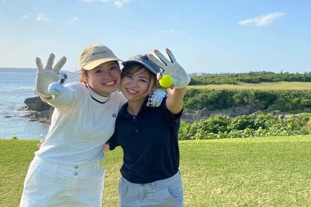 海沿いのゴルフ場で、2人の女性が笑顔で抱き合い、それぞれ白いゴルフボールと黄色いゴルフボールを手に持ってカメラに向けている。背景は青い海と緑の芝生。