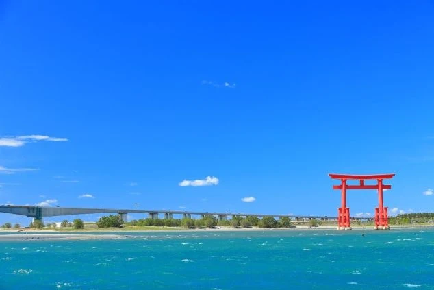 晴れた青空の下、エメラルドグリーンの海に立つ朱色の鳥居。遠景の岸辺には海を渡る長い橋が見えている。