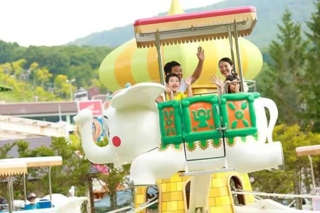 ゾウの乗り物に乗った家族が楽しそうに手を振る遊園地のアトラクション。リゾートバイトで、テーマパークやレジャー施設のスタッフとして働くイメージが伝わる画像。