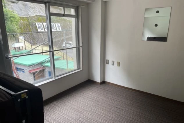 窓の外に建物とフェンスが見える部屋。壁にはコンセントと小さな鏡があり、茶色のカーペットが敷かれている。
