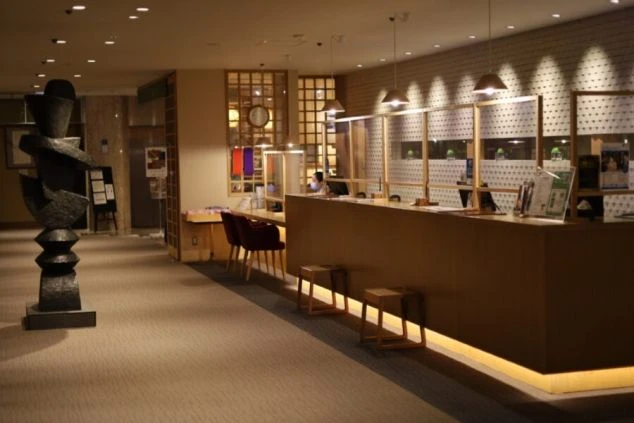 間接照明が明るいホテルロビー。木目調の受付カウンターと、現代アート風の彫刻がある。