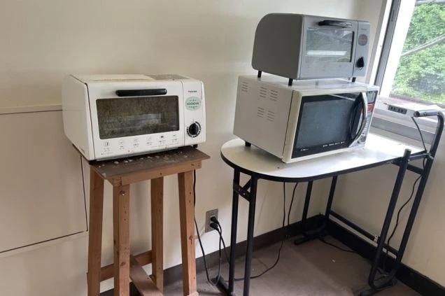 窓の近くに、トースター、電子レンジ、オーブントースターが置かれた共有キッチンの家電スペースの画像。