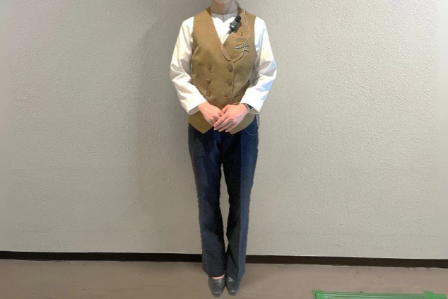 白い長袖シャツに茶色のダブルボタンベストと濃紺のパンツを着用した女性フロントスタッフの全身画像。
