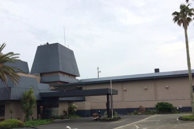 エントランスと建物外観。灰色のピラミッド型の屋根と、ベージュ色の壁、そして黒っぽい車寄せの庇（ひさし）が特徴的な建物。敷地内にはヤシの木が植えられている。