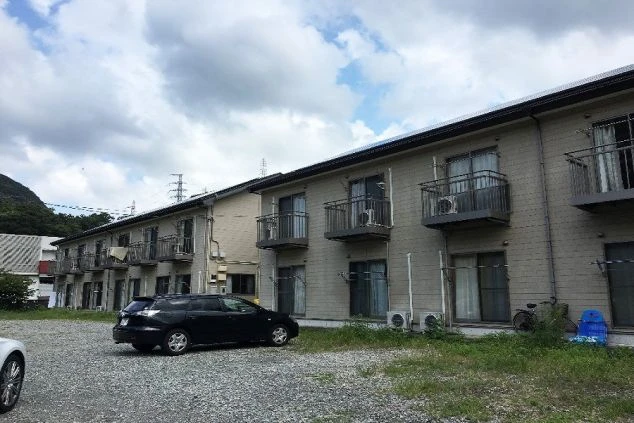 寮の外観。長い2階建ての建物で、各部屋にバルコニーがあり、駐車場は砂利敷きになっている。