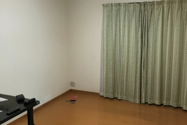 寮の居室。フローリングの床で、窓には縦縞模様のカーテンがかかっている。部屋の隅にはテーブルの一部が見える。