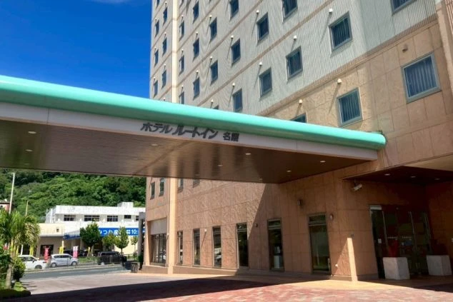 ワクトリの社員が出張で撮影した、リゾートバイトの寮の外観の画像。エントランス部分には「ホテルルートイン名護」の看板があり、青い空と南国の木々が見える。