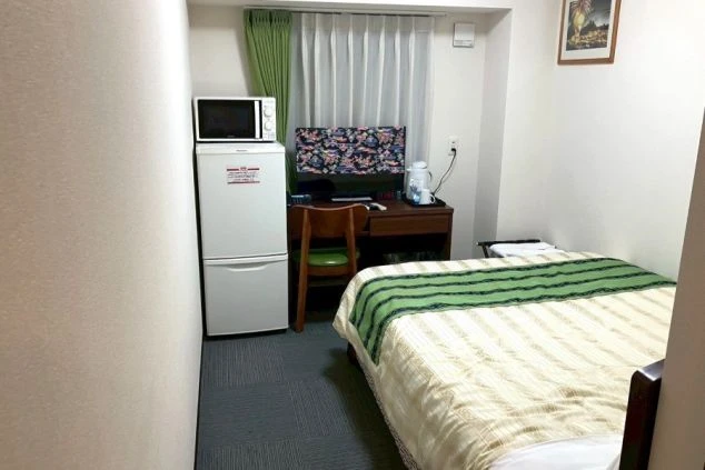 ベッド、デスク、冷蔵庫、電子レンジがある寮の部屋。ベッドには緑と白の布団が掛けられている。