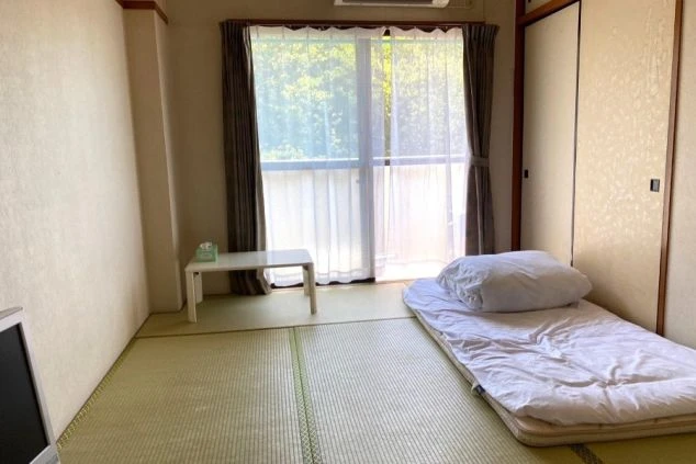 畳の上に布団が敷かれた和室の寮。窓際には小さな白いテーブルとテレビが置かれています。
