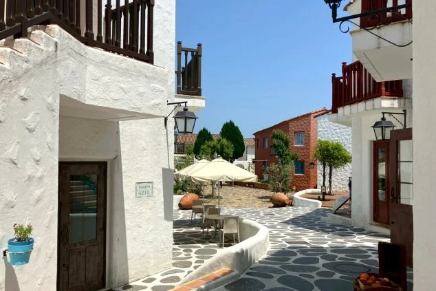 晴れた青空の下、白い漆喰壁と石積みの装飾が特徴的な、スペイン風の街並み。狭い石畳の道沿いに、赤い瓦屋根の建物が立ち並び、バルコニーや外階段が見える。建物の角には、スペインの国旗が飾られている。