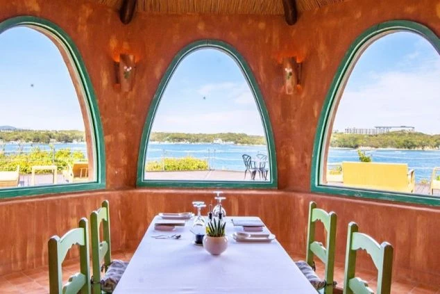 地中海風のカラフルな建物が並ぶリゾート地の広場。青と白のストライプのパラソルが設置されたカフェのテラス席があり、リゾートバイトで働く場所の魅力を伝えます。