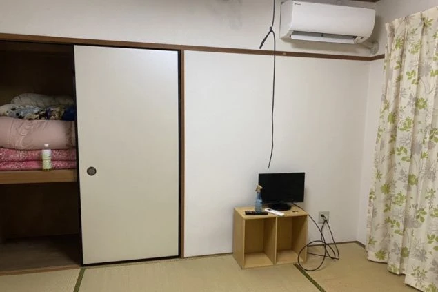 畳敷きの和室。布団が収納された押し入れ、小さなテレビ台、エアコン、花柄のカーテンがある。