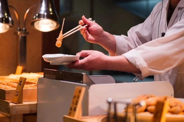 ビュッフェ会場で、白衣を着た料理人が揚げたての海老の天ぷらをお皿に取り分けている手元のアップ。