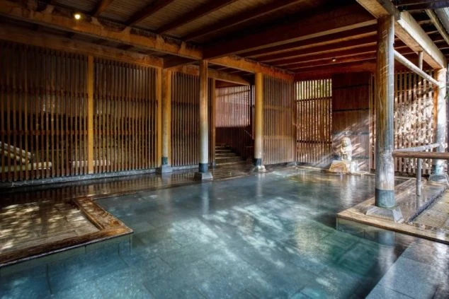 木造の柱と格子状の壁に囲まれた、落ち着いた雰囲気の広い屋内露天風呂。湯船の奥には小さな石像がある。